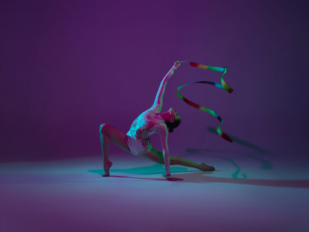 Young female athlete, rhythmic gymnastics artist on purple background with neon light. Beautiful girl practicing with equipment. Grace in performance.